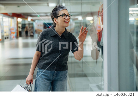 Middle-aged woman with short gray hair wearing glasses and casual clothing smiles while looking through a glass window in a shopping mall with bright interior lighting 140036321