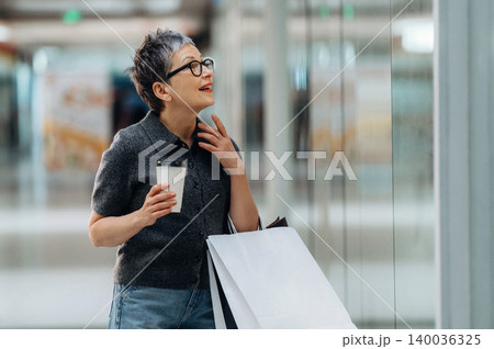 Middle-aged Asian woman with short hair holding a coffee cup and shopping bag, smiling while looking at a store window in a modern shopping mall 140036325