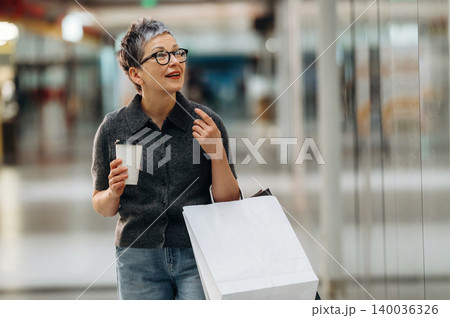 Middle-aged woman with short gray hair holds coffee cup and shopping bag while walking through a modern shopping mall with bright lighting and glass walls 140036326