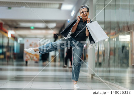 Senior woman with glasses joyfully poses in a shopping mall while holding shopping bags, showcasing a vibrant retail environment with reflective surfaces Senior woman with glasses joyfully poses in a shopping mall while holding shopping bags, showcasing a vibrant retail environment with reflective surfaces 140036338