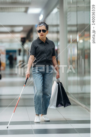 Elderly woman with short gray hair wearing sunglasses and a black shirt walks through a shopping mall holding shopping bags and using a white cane for navigation Elderly woman with short gray hair wearing sunglasses and a black shirt walks through a shopping mall holding shopping bags and using a white cane for navigation 140036339