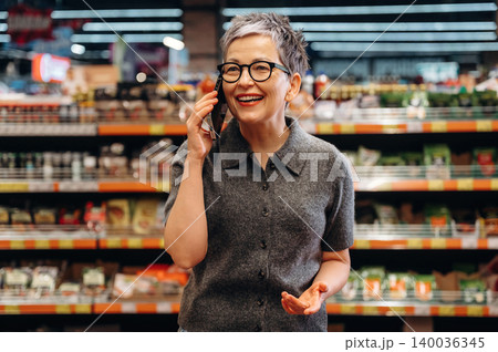 Senior woman with short gray hair wearing glasses talks on a smartphone while smiling in a grocery store aisle filled with various food products and shelves 140036345