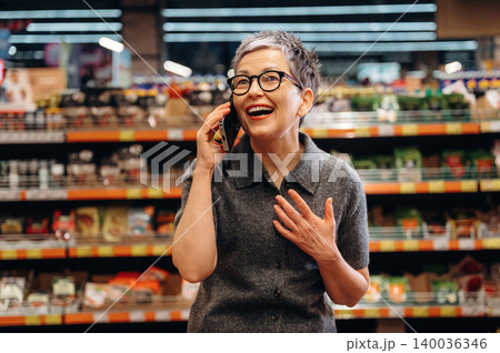 Middle-aged woman with short gray hair wearing glasses talks on a smartphone while smiling inside a grocery store with shelves of products in the background 140036346