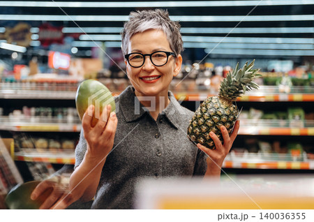 Mature woman with short gray hair holding a mango and pineapple in a grocery store, colorful shelves filled with fruits and vegetables visible in the background Mature woman with short gray hair holding a mango and pineapple in a grocery store, colorful shelves filled with fruits and vegetables visible in the background 140036355