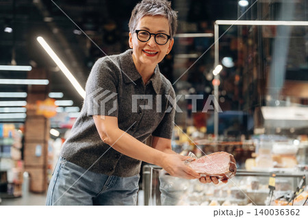 Mature Asian woman with short gray hair wearing glasses holds a piece of meat in a grocery store, showcasing fresh food in a well-lit market environment Mature Asian woman with short gray hair wearing glasses holds a piece of meat in a grocery store, showcasing fresh food in a well-lit market environment 140036362