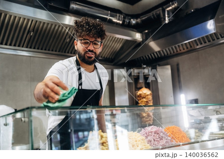 Male chef with curly hair wearing glasses cleans glass display case in a modern restaurant kitchen, various fresh ingredients visible in the background Male chef with curly hair wearing glasses cleans glass display case in a modern restaurant kitchen, various fresh ingredients visible in the background 140036562