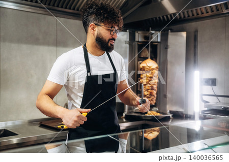 Male chef with curly hair wearing glasses and black apron holds a knife while preparing food in a modern kitchen with a vertical rotisserie in the background Male chef with curly hair wearing glasses and black apron holds a knife while preparing food in a modern kitchen with a vertical rotisserie in the background 140036565