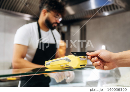 Male cashier wearing black apron processes payment with a yellow card reader as a customer presents a black credit card at a modern cafe counter Male cashier wearing black apron processes payment with a yellow card reader as a customer presents a black credit card at a modern cafe counter 140036590