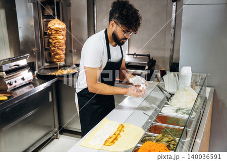 Male chef in black apron prepares a wrap with various ingredients on a countertop in a modern kitchen with a vertical rotisserie in the background Male chef in black apron prepares a wrap with various ingredients on a countertop in a modern kitchen with a vertical rotisserie in the background 140036591