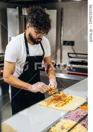 Male chef with curly hair prepares a taco with various toppings at a food counter in a modern kitchen, equipped with cooking appliances and fresh ingredients 140036596