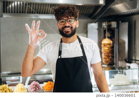 Male chef wearing glasses and gloves shows an okay hand gesture while standing in front of a food counter with various ingredients in a restaurant kitchen Male chef wearing glasses and gloves shows an okay hand gesture while standing in front of a food counter with various ingredients in a restaurant kitchen 140036601