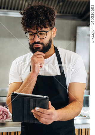 Male chef with curly hair and beard wearing black apron checks recipe on tablet while standing in modern kitchen with stainless steel appliances and ingredients visible 140036603