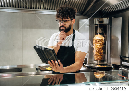 Male chef in black apron using tablet while standing in modern kitchen with vertical rotisserie and glass counter displaying food preparation area Male chef in black apron using tablet while standing in modern kitchen with vertical rotisserie and glass counter displaying food preparation area 140036619