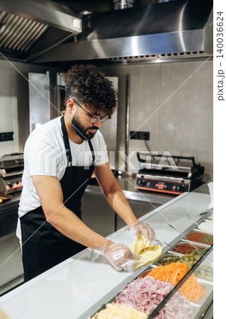 Male chef with curly hair wearing glasses prepares fresh ingredients in a modern kitchen, arranging vegetables and sauces on a countertop for meal preparation 140036624