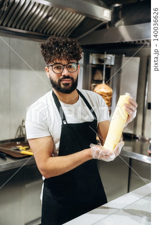 Male chef wearing glasses and black apron holds a rolled tortilla in a modern kitchen with cooking equipment and vertical grill in the background 140036626