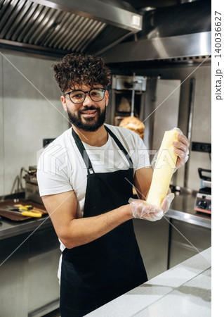 Male chef with curly hair wearing glasses and black apron holds a rolled pastry in a modern kitchen with stainless steel appliances and cooking equipment visible 140036627
