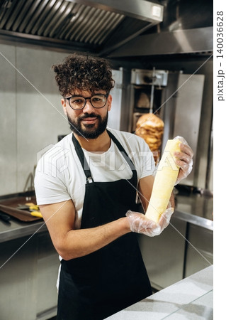 Male chef with curly hair wearing glasses holds a rolled tortilla in a commercial kitchen with a vertical rotisserie in the background 140036628