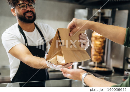 Male food vendor in black apron hands over a takeout bag to a customer at a fast food counter with a vertical rotisserie in the background Male food vendor in black apron hands over a takeout bag to a customer at a fast food counter with a vertical rotisserie in the background 140036633