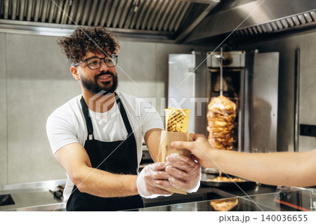 Male food vendor in black apron hands over wrapped kebab to customer at counter in modern fast-food restaurant with vertical grill in background Male food vendor in black apron hands over wrapped kebab to customer at counter in modern fast-food restaurant with vertical grill in background 140036655