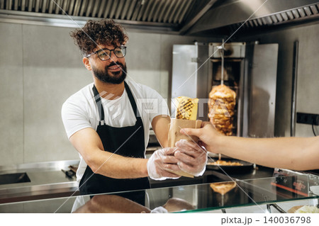 Male food vendor with curly hair in black apron hands over wrapped kebab to customer at counter in modern fast food restaurant with vertical grill in background Male food vendor with curly hair in black apron hands over wrapped kebab to customer at counter in modern fast food restaurant with vertical grill in background 140036798
