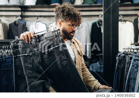 Male customer with curly hair examines black jeans while shopping in a clothing store, racks of apparel and merchandise visible in the background 140036802