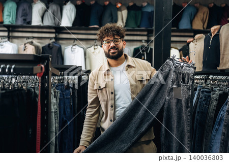 Male customer in a clothing store holds up a pair of dark jeans while browsing through a rack of apparel, with various shirts displayed in the background 140036803
