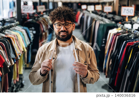 Male tailor with curly hair and glasses holds measuring tape while standing in a clothing store aisle filled with colorful garments on hangers 140036816
