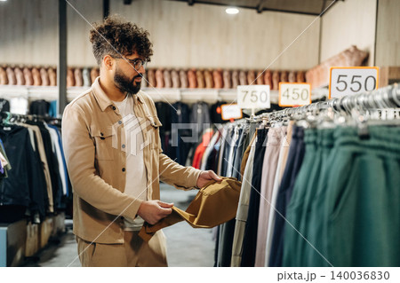 Male shopper with curly hair examines clothing items in a retail store, surrounded by racks of apparel and price tags visible in a well-lit indoor environment 140036830