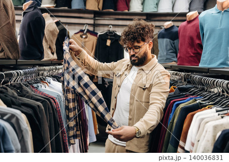 Man with curly hair examines plaid shirt while shopping for clothing in a retail store, surrounded by various garments on hangers in the background Man with curly hair examines plaid shirt while shopping for clothing in a retail store, surrounded by various garments on hangers in the background 140036831