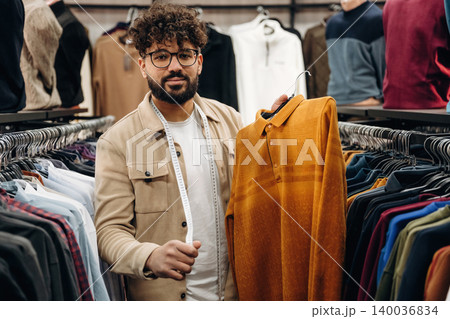 Male fashion consultant holding orange polo shirt while standing in clothing store surrounded by various garments on hangers in the background 140036834