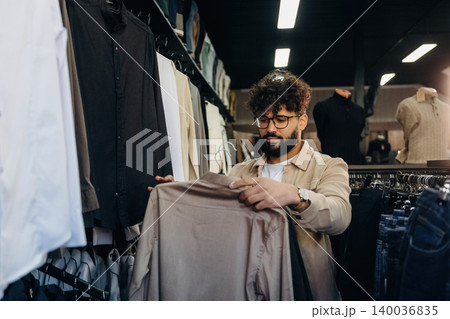 Male shopper with curly hair examines beige shirt while browsing clothing rack in modern retail store with various garments displayed in the background 140036835