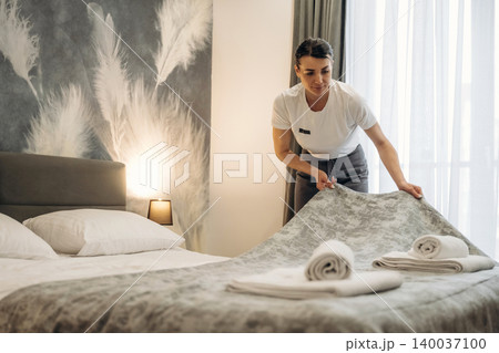 Hotel housekeeper arranging bedding in a modern guest room with decorative wall and neatly folded towels on the bed, natural light streaming through the window Hotel housekeeper arranging bedding in a modern guest room with decorative wall and neatly folded towels on the bed, natural light streaming through the window 140037100