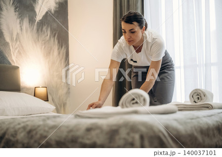 Hotel housekeeping staff member arranging towels on a neatly made bed in a modern guest room with soft lighting and elegant decor elements Hotel housekeeping staff member arranging towels on a neatly made bed in a modern guest room with soft lighting and elegant decor elements 140037101