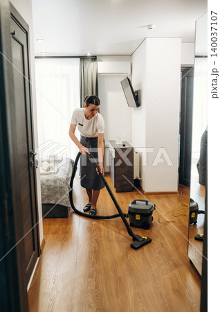 Female hotel staff member vacuuming wooden floor in a modern hotel room with a bed, mirror, and television visible in the background Female hotel staff member vacuuming wooden floor in a modern hotel room with a bed, mirror, and television visible in the background 140037107