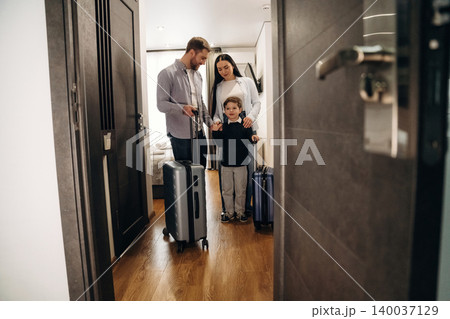 Family of three with luggage standing in hotel room doorway, modern decor visible, wooden floor, and soft lighting creating a welcoming atmosphere 140037129