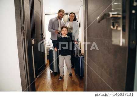 Family with luggage entering hotel room, young boy leading with suitcase, parents following, modern interior design visible in the background 140037131