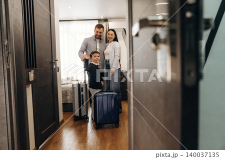 Family with luggage standing in hotel room doorway, smiling together as they prepare for their stay in a modern accommodation with stylish decor and wooden flooring Family with luggage standing in hotel room doorway, smiling together as they prepare for their stay in a modern accommodation with stylish decor and wooden flooring 140037135