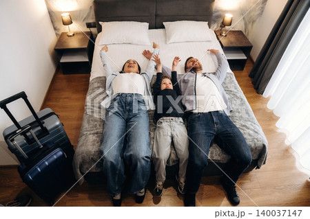 Family of three lying on a hotel bed with arms raised, suitcase nearby, in a modern room with wooden flooring and soft lighting, creating a cozy atmosphere Family of three lying on a hotel bed with arms raised, suitcase nearby, in a modern room with wooden flooring and soft lighting, creating a cozy atmosphere 140037147