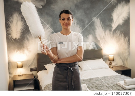 Hotel housekeeper standing in a well-decorated guest room, holding a feather duster with a modern design, showcasing a clean and inviting atmosphere Hotel housekeeper standing in a well-decorated guest room, holding a feather duster with a modern design, showcasing a clean and inviting atmosphere 140037307