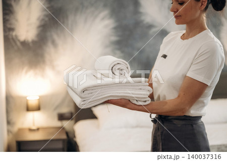 Female hotel staff member in a white shirt holds neatly stacked towels in a modern hotel room with a stylish decor and a bedside lamp illuminated in the background Female hotel staff member in a white shirt holds neatly stacked towels in a modern hotel room with a stylish decor and a bedside lamp illuminated in the background 140037316
