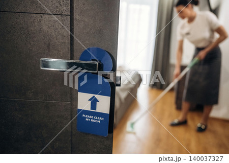 Hotel room door with a "Please Clean My Room" sign and a housekeeper cleaning the floor with a mop in the background, showcasing a tidy interior environment Hotel room door with a "Please Clean My Room" sign and a housekeeper cleaning the floor with a mop in the background, showcasing a tidy interior environment 140037327