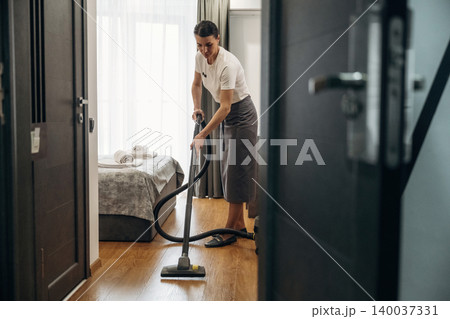 Hotel housekeeping staff member vacuuming wooden floor in a guest room, neatly made bed with towels visible in the background, bright natural light streaming through the window Hotel housekeeping staff member vacuuming wooden floor in a guest room, neatly made bed with towels visible in the background, bright natural light streaming through the window 140037331