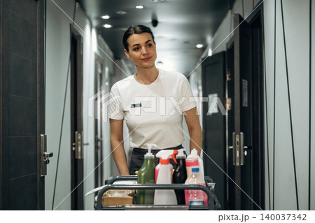 Hotel housekeeping staff member pushing a cart filled with cleaning supplies down a corridor with closed doors and modern lighting fixtures visible in the background Hotel housekeeping staff member pushing a cart filled with cleaning supplies down a corridor with closed doors and modern lighting fixtures visible in the background 140037342