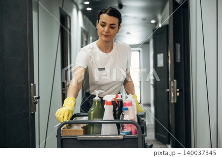 Female hotel housekeeper in white shirt and yellow gloves pushes cleaning cart filled with supplies down a modern hotel corridor with doors and lighting visible Female hotel housekeeper in white shirt and yellow gloves pushes cleaning cart filled with supplies down a modern hotel corridor with doors and lighting visible 140037345