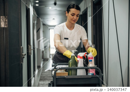 Hotel housekeeping staff member in uniform pushes cleaning cart filled with supplies down a corridor, with doors and modern lighting visible in the background Hotel housekeeping staff member in uniform pushes cleaning cart filled with supplies down a corridor, with doors and modern lighting visible in the background 140037347