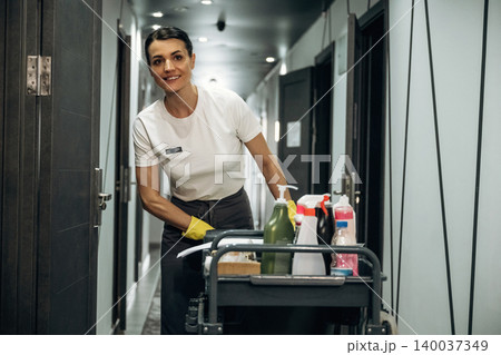 Hotel housekeeping staff member wearing uniform smiles while pushing a cleaning cart filled with supplies in a corridor with guest room doors visible Hotel housekeeping staff member wearing uniform smiles while pushing a cleaning cart filled with supplies in a corridor with guest room doors visible 140037349