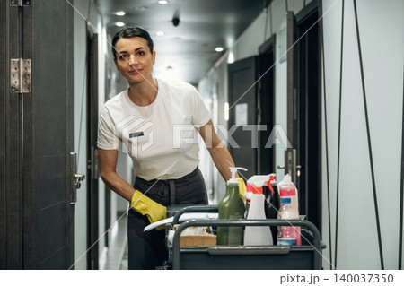 Hotel housekeeping staff member wearing gloves pushes a cleaning cart filled with supplies along a corridor with doors and modern lighting in the background 140037350