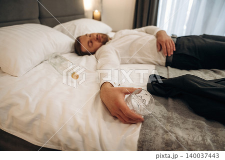 Male individual in white shirt lying on a hotel bed with an empty bottle and glass nearby, soft lighting creating a relaxed atmosphere in the room 140037443