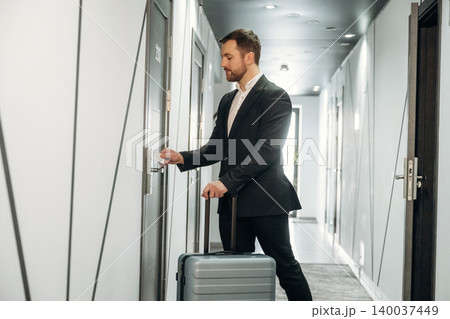 Businessman in a black suit standing in hotel corridor, holding a suitcase and preparing to open room door with keycard, modern interior design visible Businessman in a black suit standing in hotel corridor, holding a suitcase and preparing to open room door with keycard, modern interior design visible 140037449