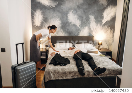 Hotel room scene with a female staff member assisting a male guest lying on the bed, featuring modern decor and a suitcase nearby Hotel room scene with a female staff member assisting a male guest lying on the bed, featuring modern decor and a suitcase nearby 140037480